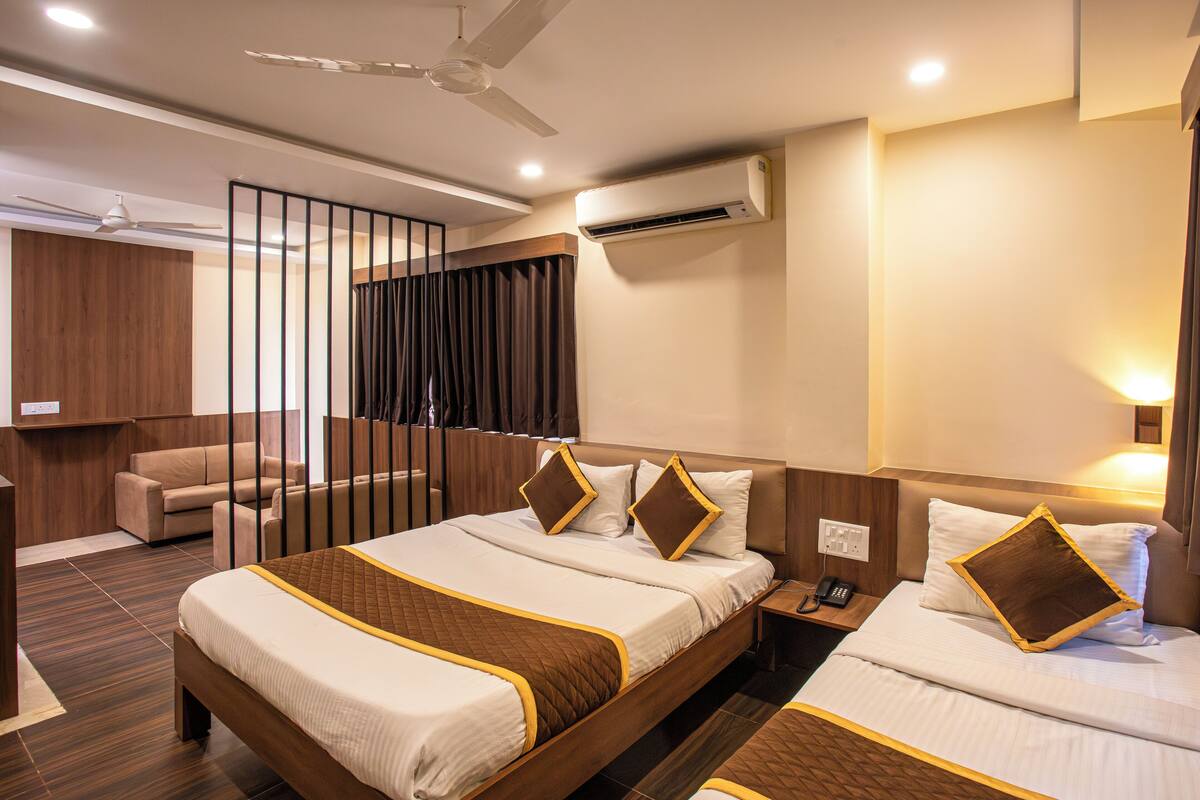A hotel room with a large bed, a smaller bed, a desk, a chair, a sofa, a curtain, a ceiling fan, and an air conditioning unit.