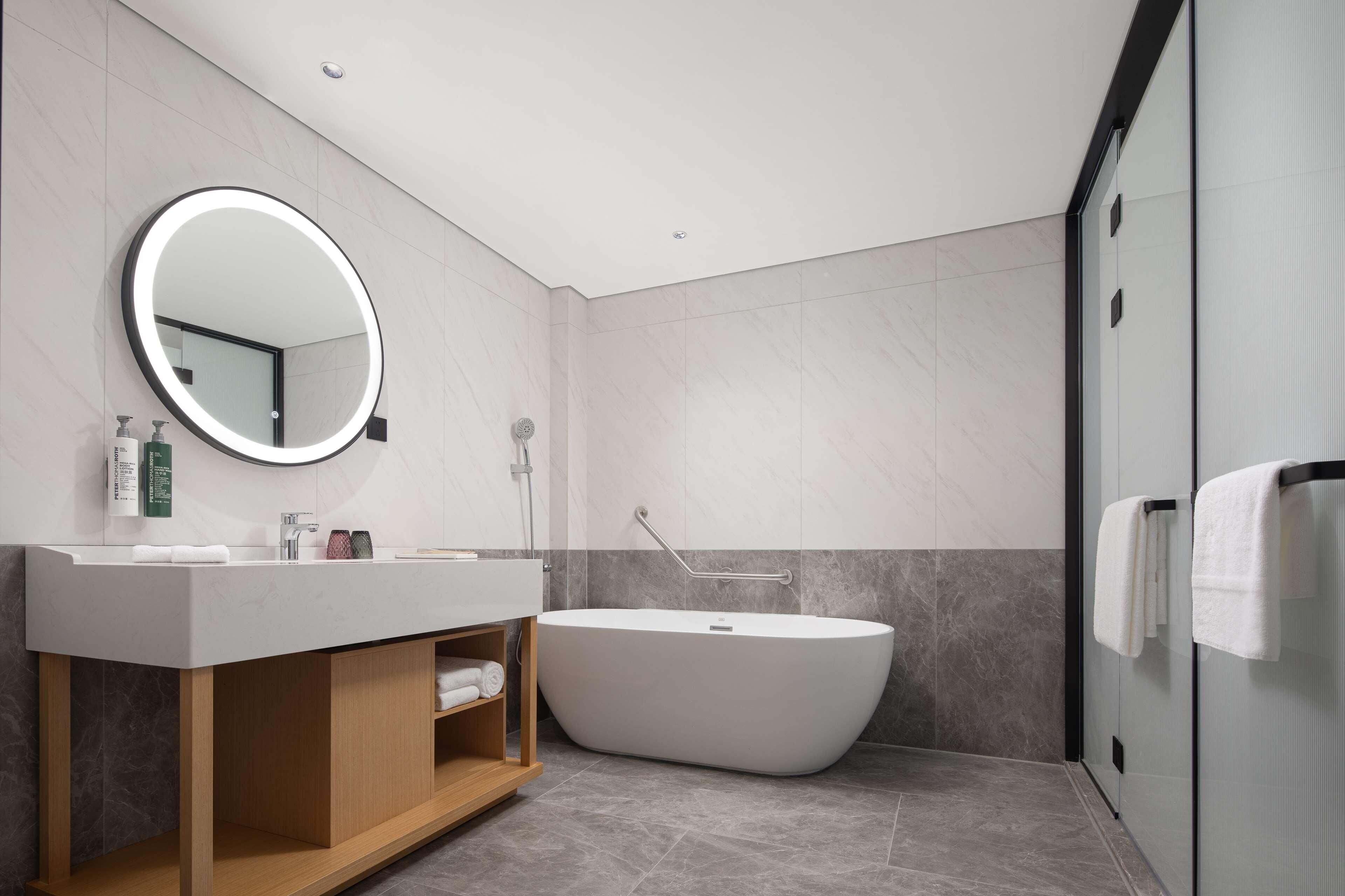 A modern bathroom with a freestanding bathtub, a round mirror with a light, and a vanity with a wooden cabinet.