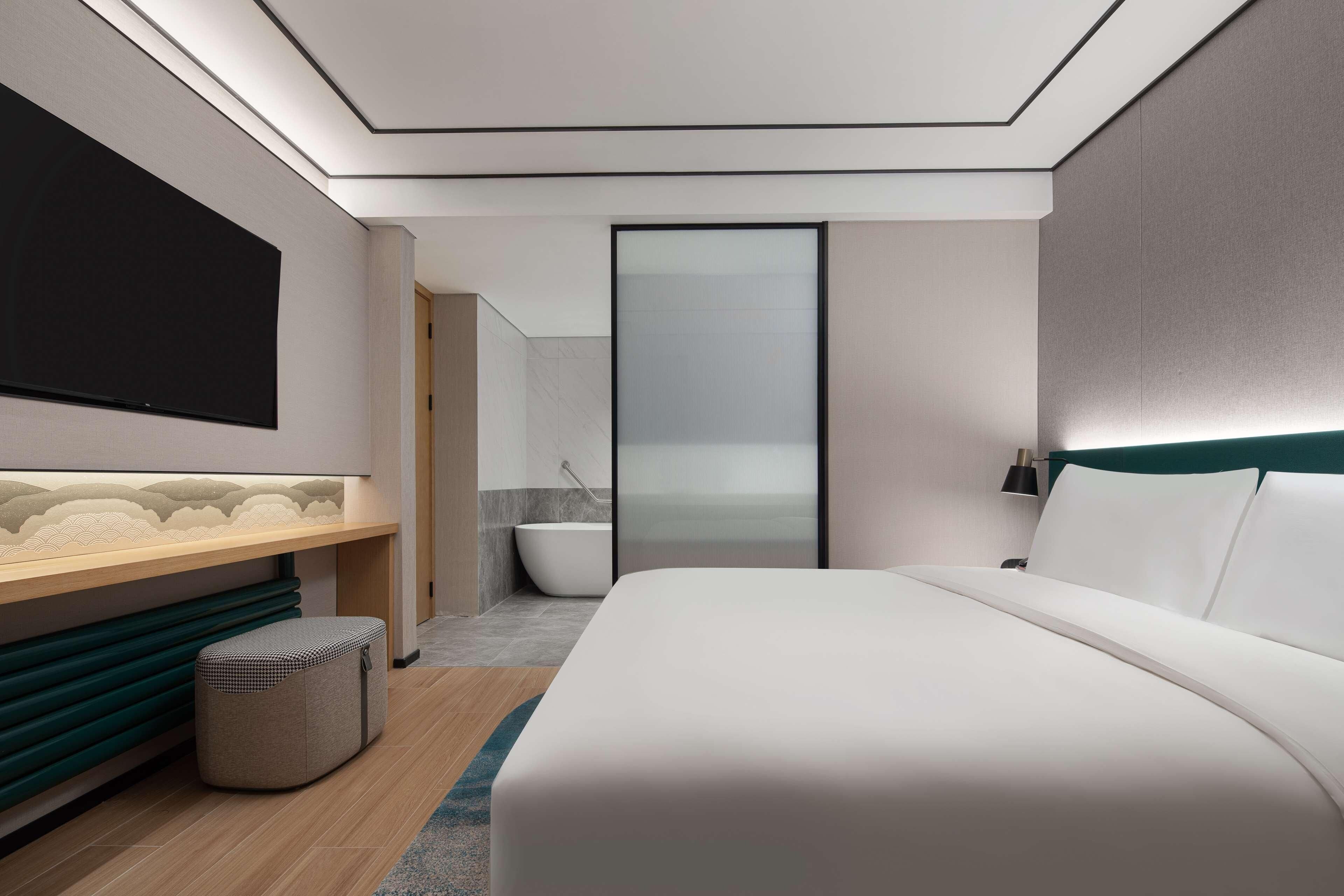 A modern hotel room with a large bed, a vanity with a mirror, a bathtub, and a view of the sea through a glass door.