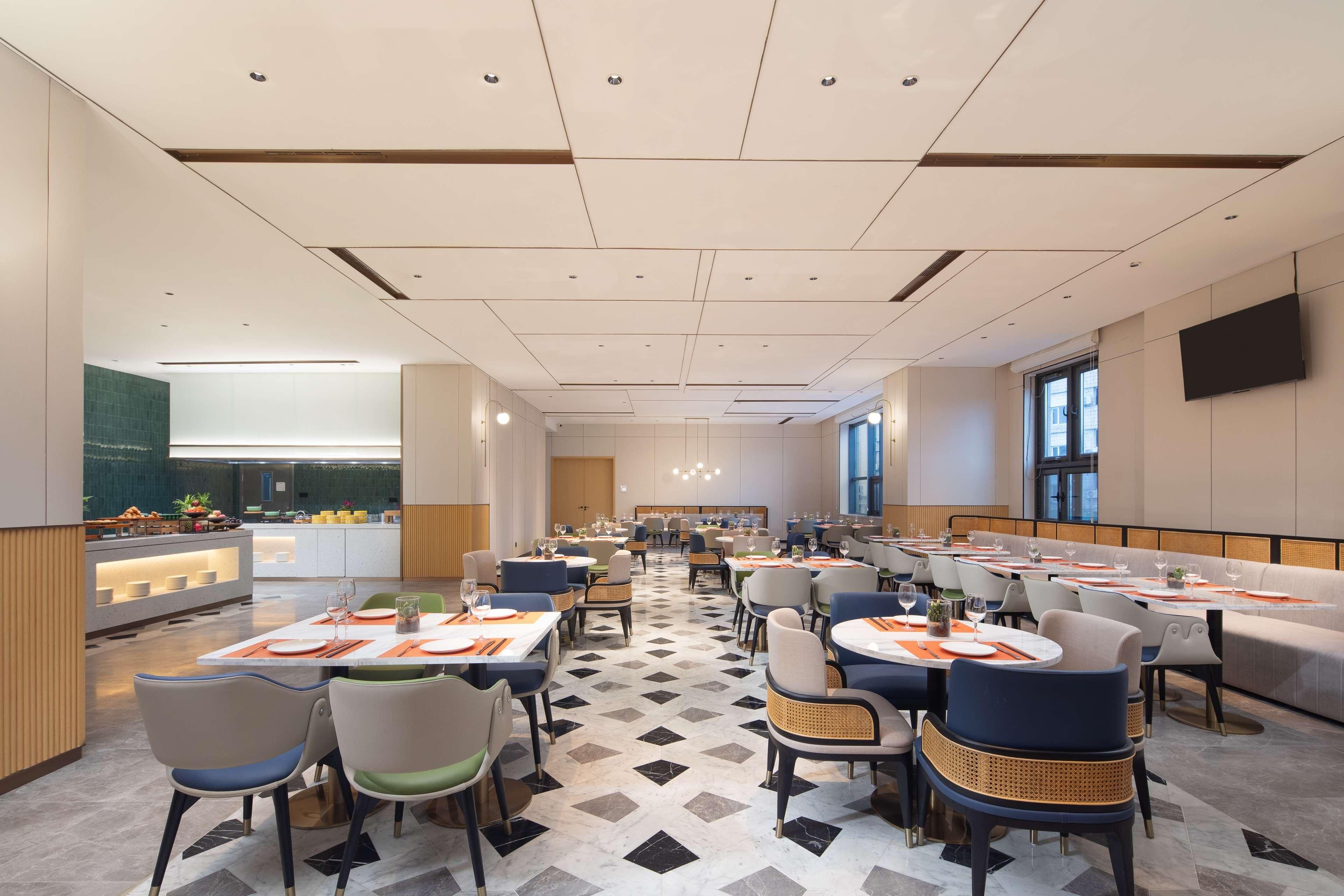 A spacious dining area with multiple tables and chairs, a kitchen in the background, and a large window providing natural light.