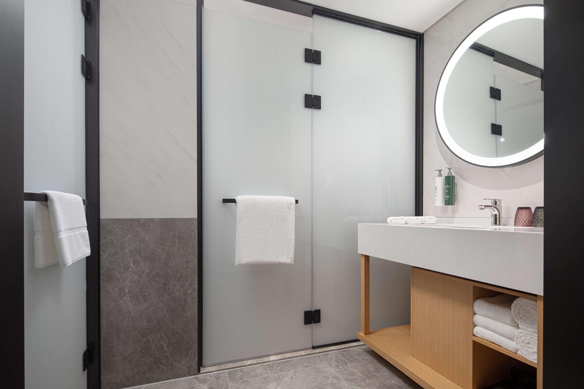 Un baño moderno con mampara de vidrio, espejo redondo, lavamanos blanco y toallas ordenadas.
