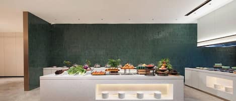 A modern hotel lobby with a central buffet area featuring various food items and a seating area with a geometric patterned floor.