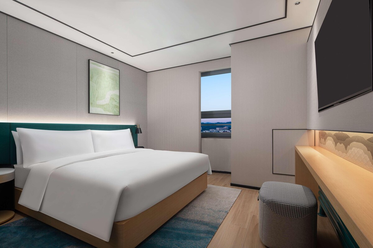 A modern hotel room with a bed, a small stool, a window with a view, and a wall-mounted TV.