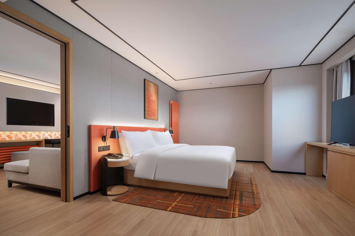 Una habitación de hotel moderna con cama, mesita de noche, lámpara, televisor y un pequeño rincón de estar.