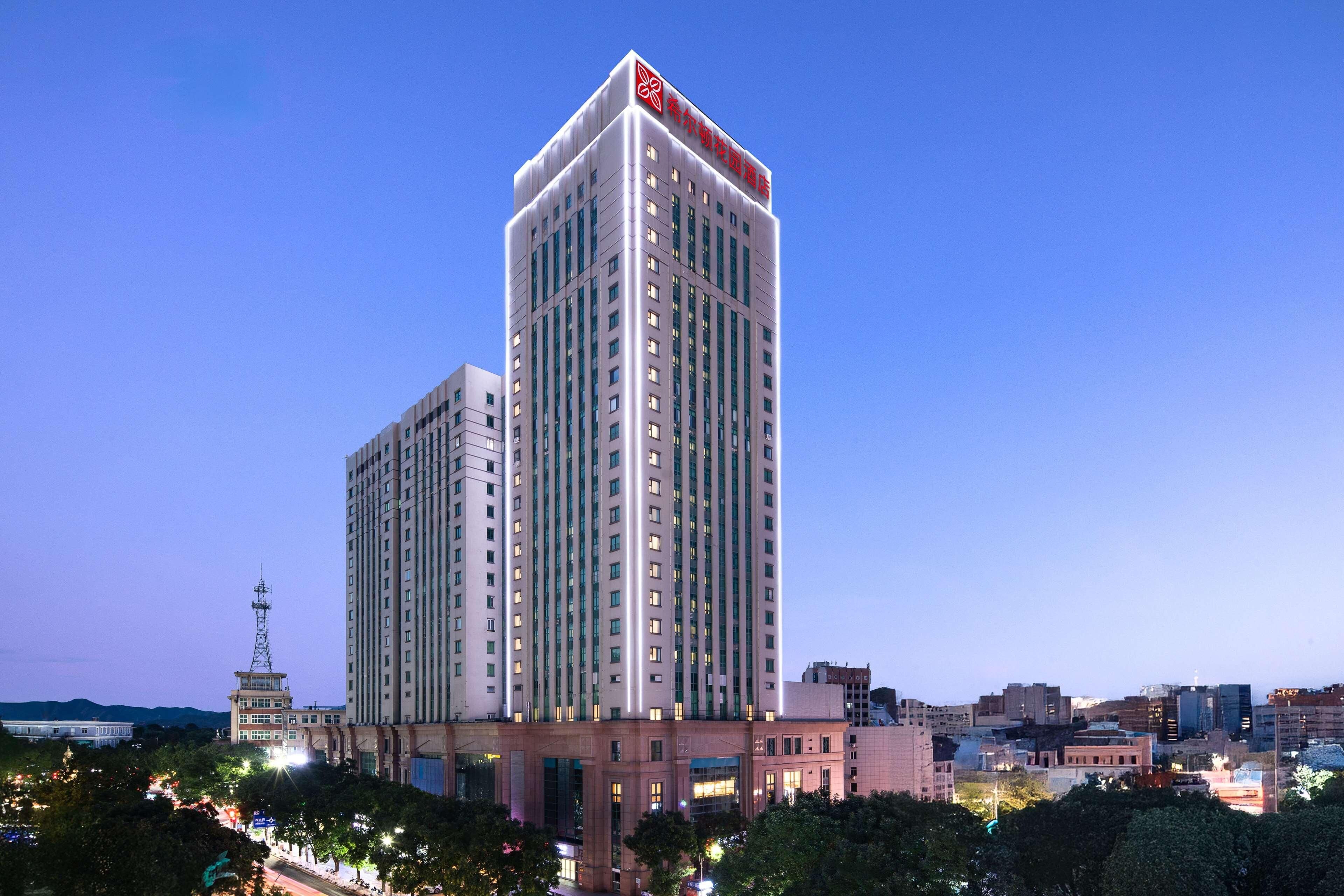 A high-rise hotel with a prominent red logo on the top, surrounded by other buildings and trees.