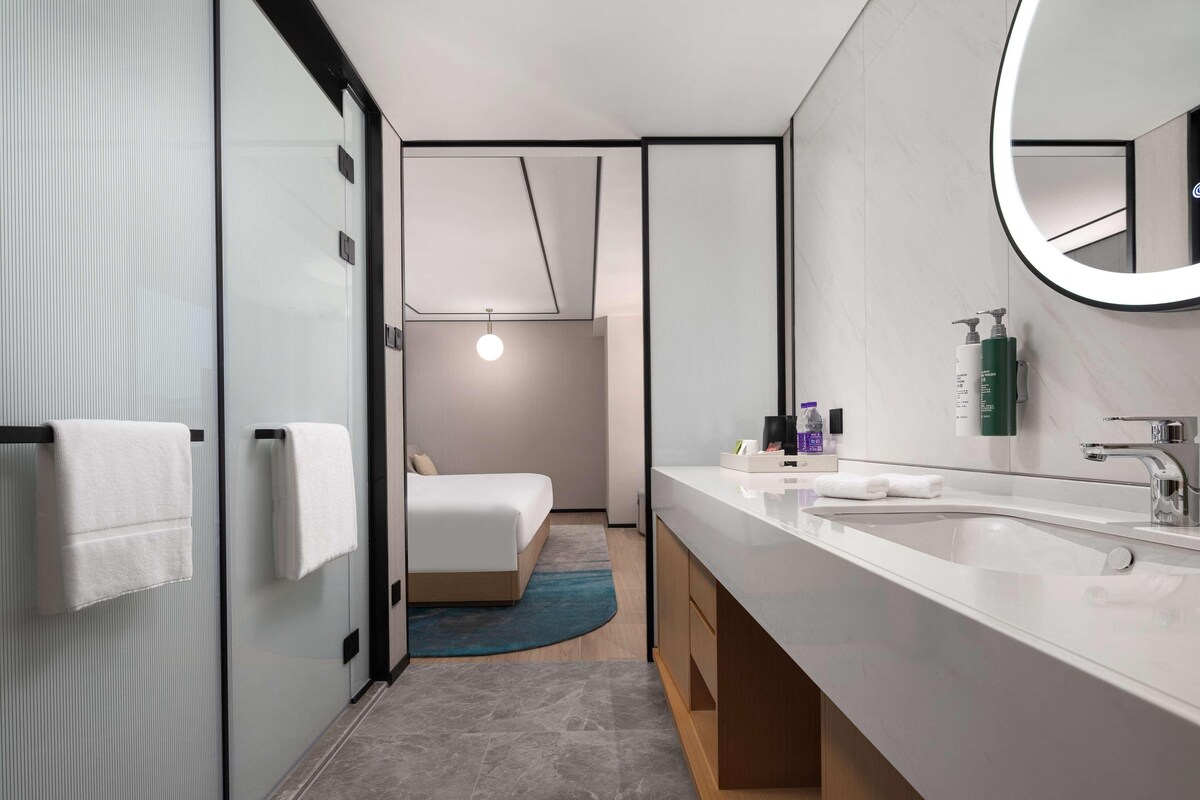 A modern hotel bathroom with a large mirror, two sinks, and a bed visible through a glass partition.