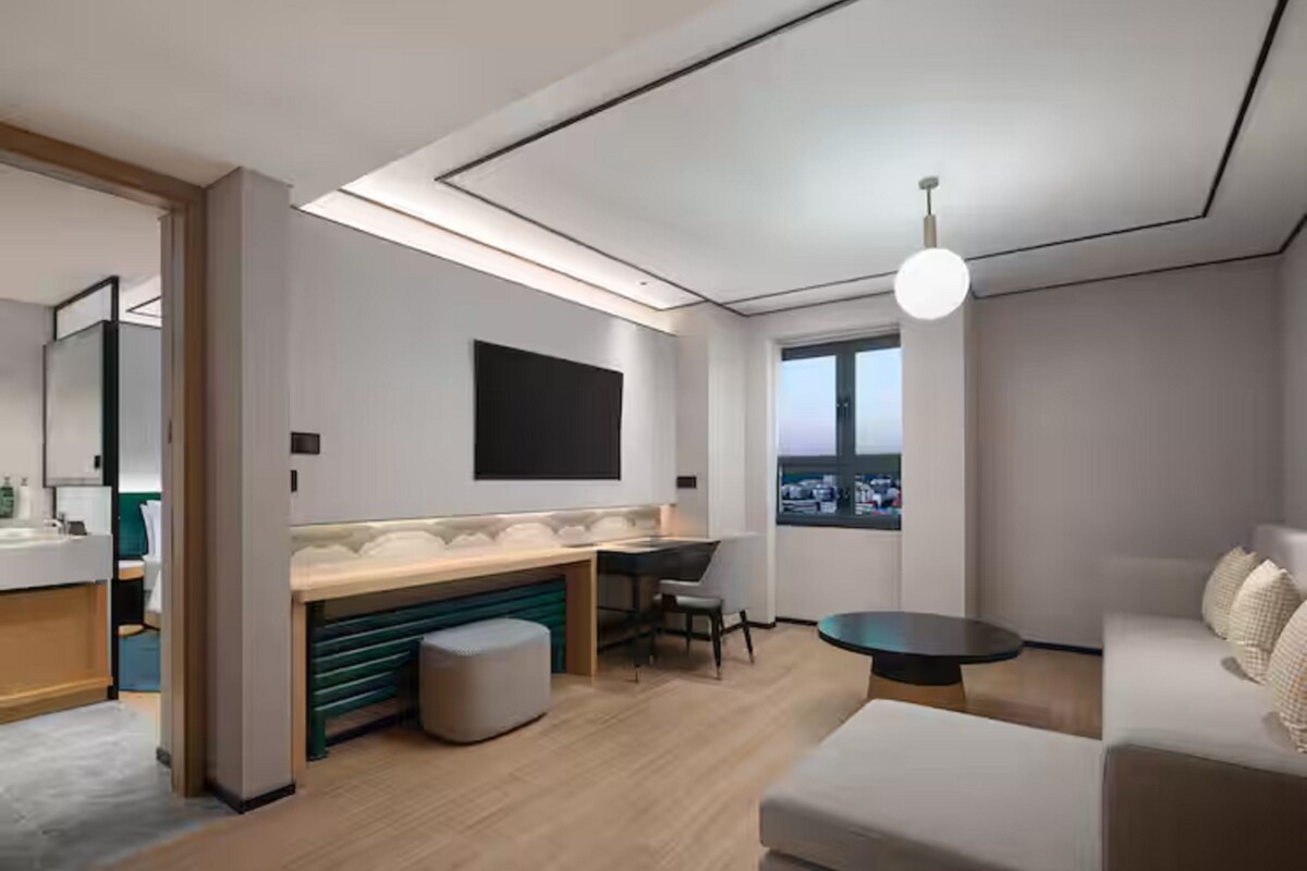 A modern hotel room with a kitchenette, a large window, and a seating area.