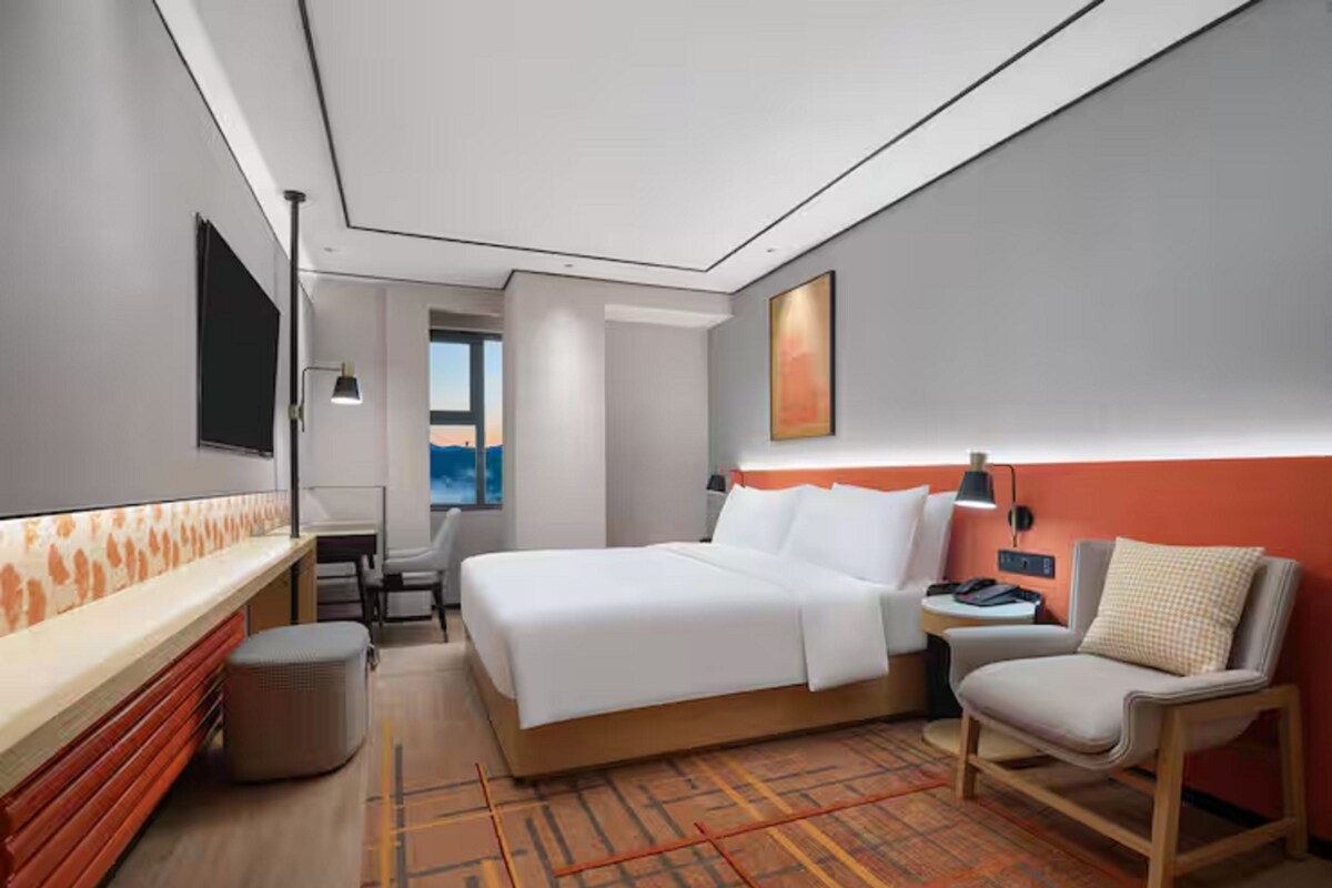 A modern hotel room with a bed, a desk, a chair, and a view of the outdoors.