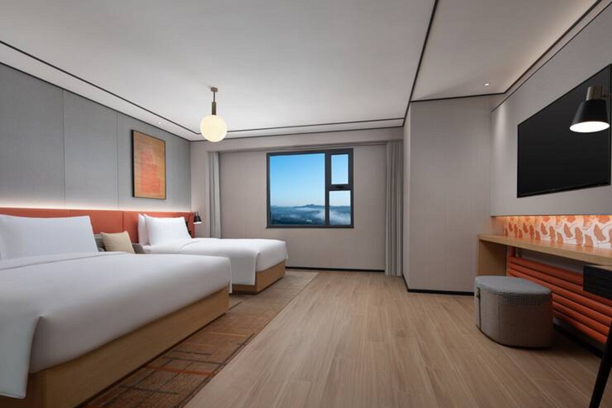 Una habitación de hotel moderna con dos camas, un ventanal grande, un televisor y un pequeño rincón de descanso.