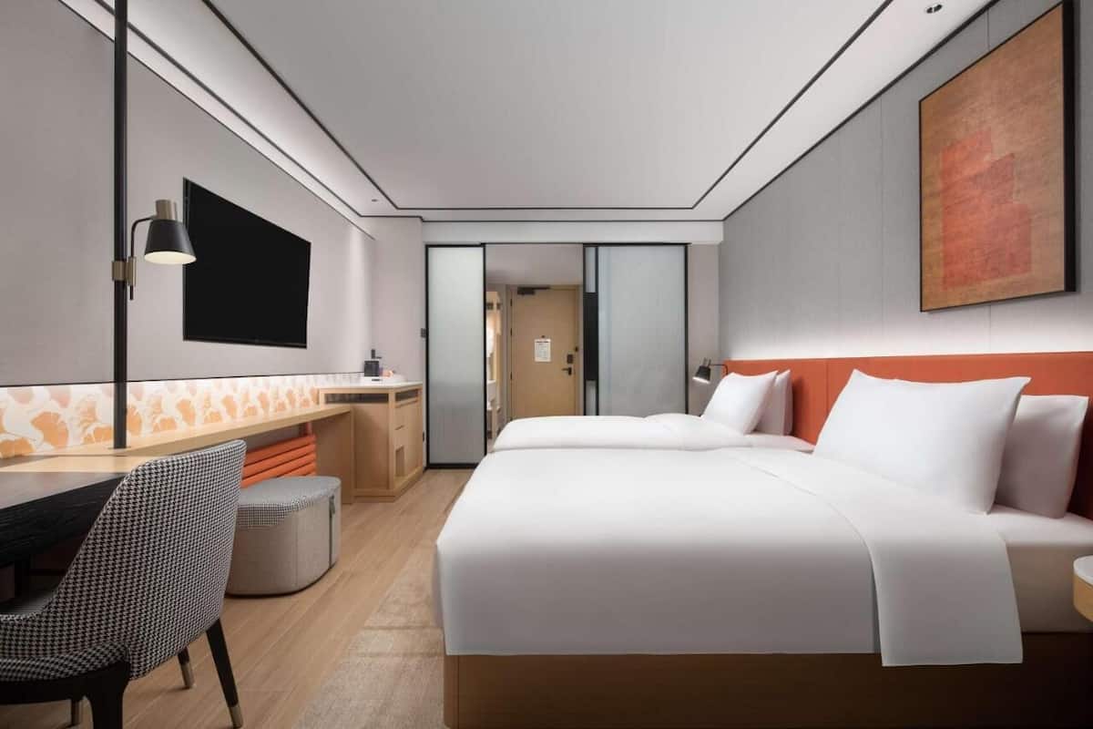 Ein modernes Hotelzimmer mit einem großen Bett, einem Schreibtisch und einem Sessel. Ein Fernseher ist an der Wand angebracht und über dem Bett hängt ein Gemälde.