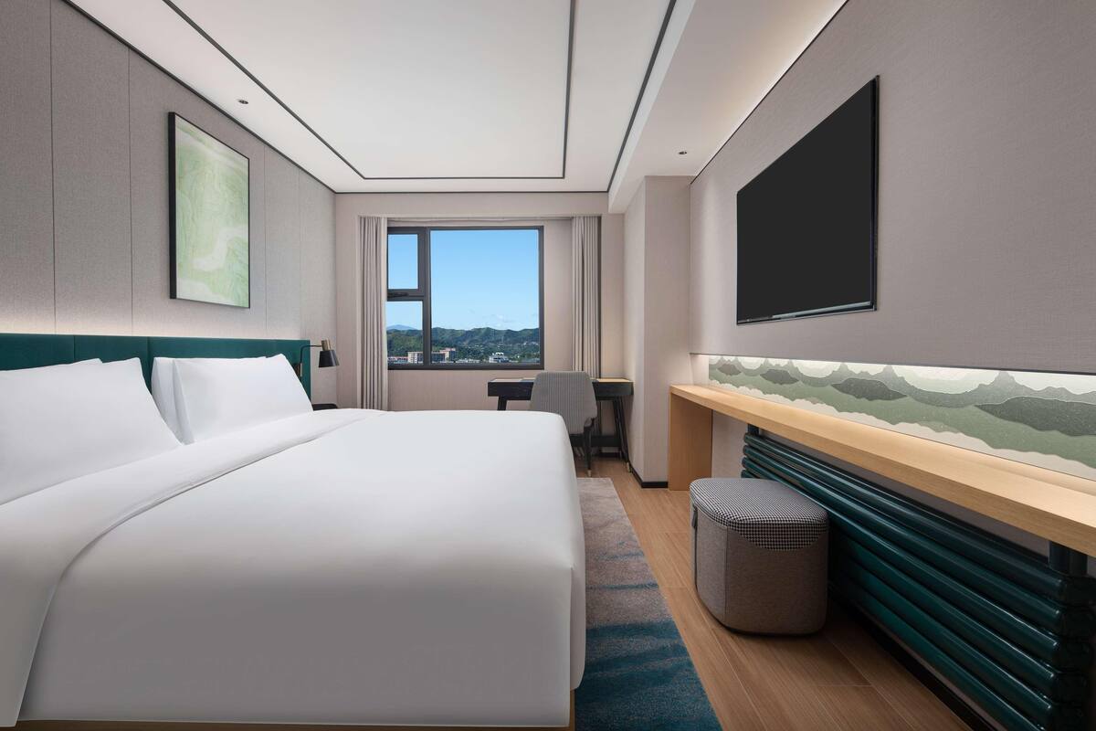 Ein modernes Hotelzimmer mit einem großen Bett, einem Schreibtisch, einem Sessel, einem Fernseher und Blick auf die Stadt.