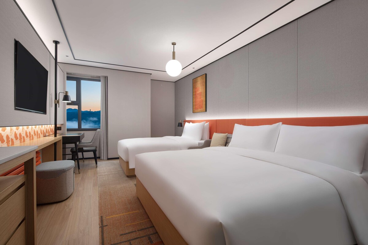 A modern hotel room with two beds, a desk, a chair, and a large window offering a view of the mountains.