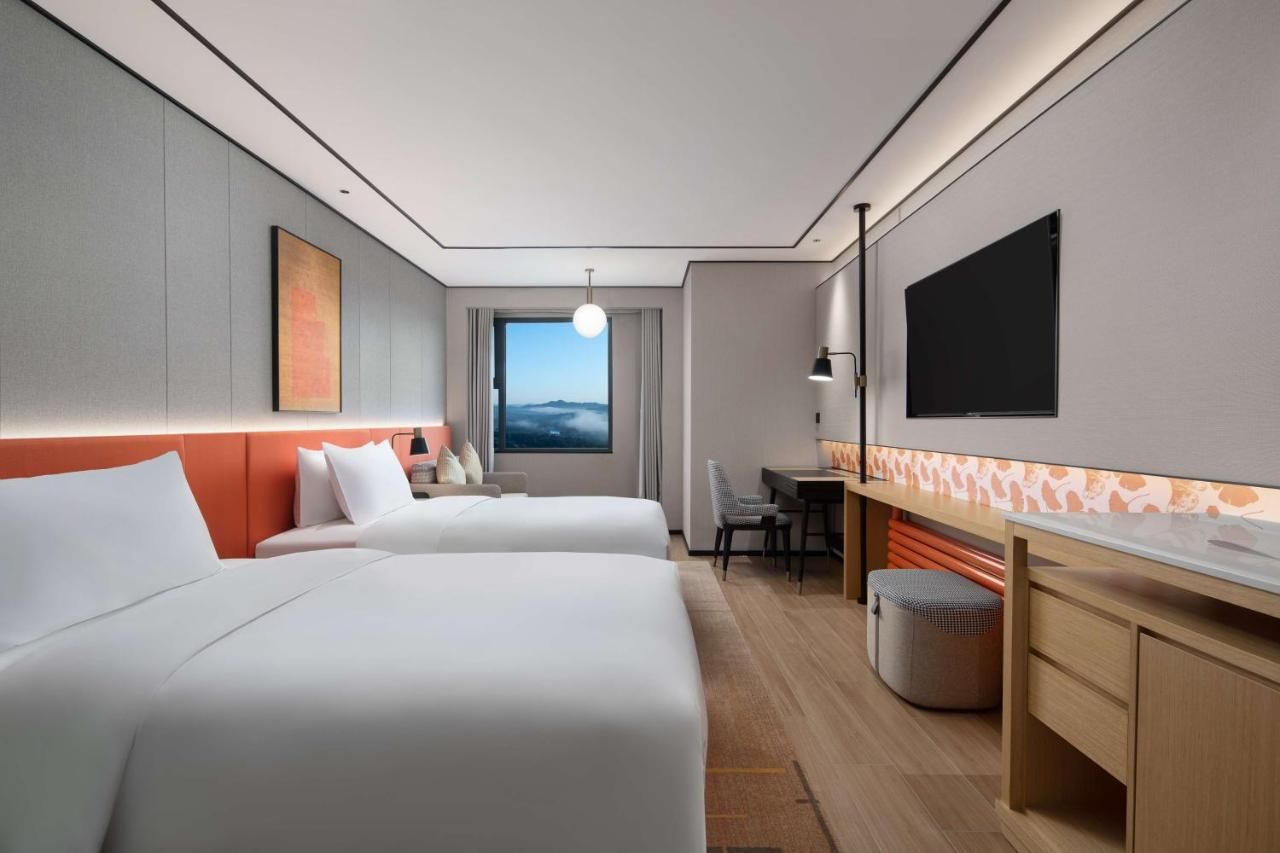 A modern hotel room with two beds, a desk, a TV, and a view of the sky.