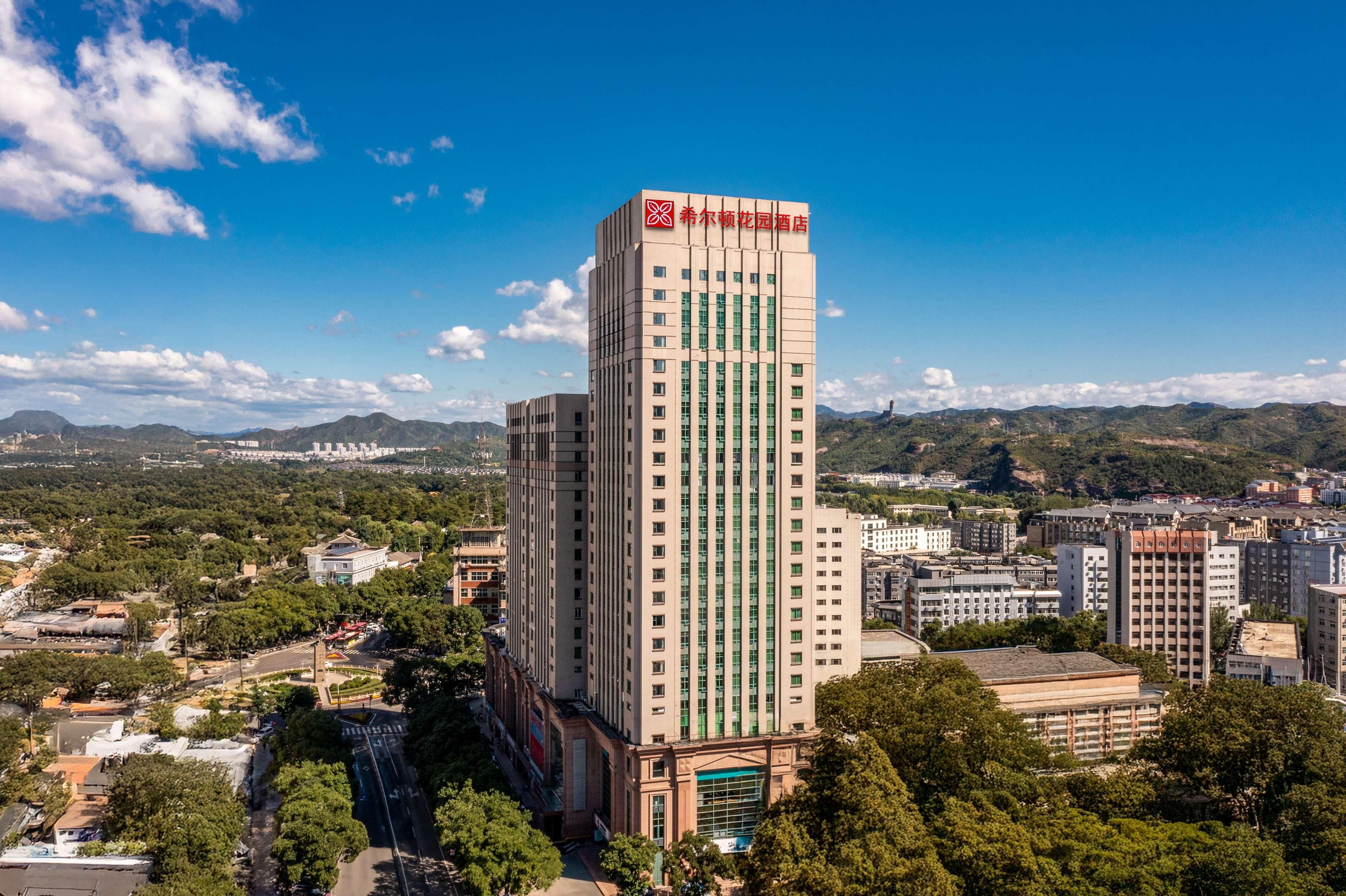 A tall hotel with a prominent red logo on the top, surrounded by urban buildings and greenery.