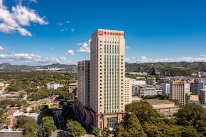 A tall hotel with a prominent red logo on the top, surrounded by urban buildings and greenery.
