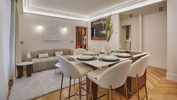 Un'area pranzo moderna con un tavolo rettangolare apparecchiato per quattro persone, circondato da sedie abbinate. Sullo sfondo, si intravede un divano e un tavolino.