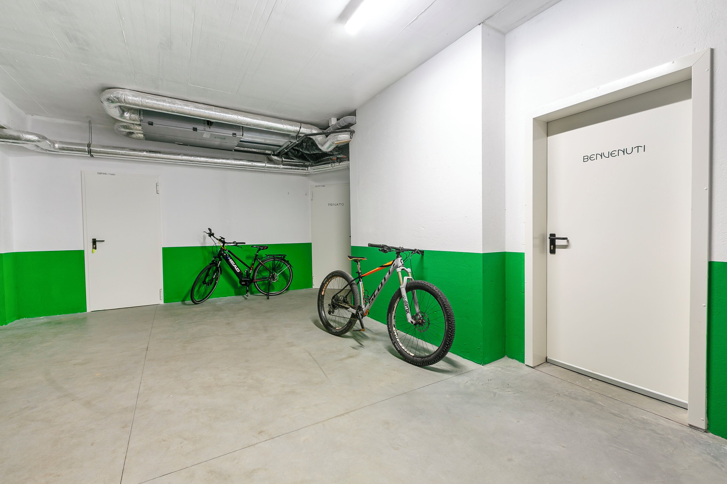 A clean, well-lit indoor space with a green and white color scheme, featuring a door labeled "BENJENUTI" and two bicycles parked against the wall.