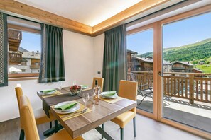 Un comedor con una mesa preparada para cuatro, vistas a un paisaje montañoso a través de amplios ventanales y un balcón con barandilla de madera.