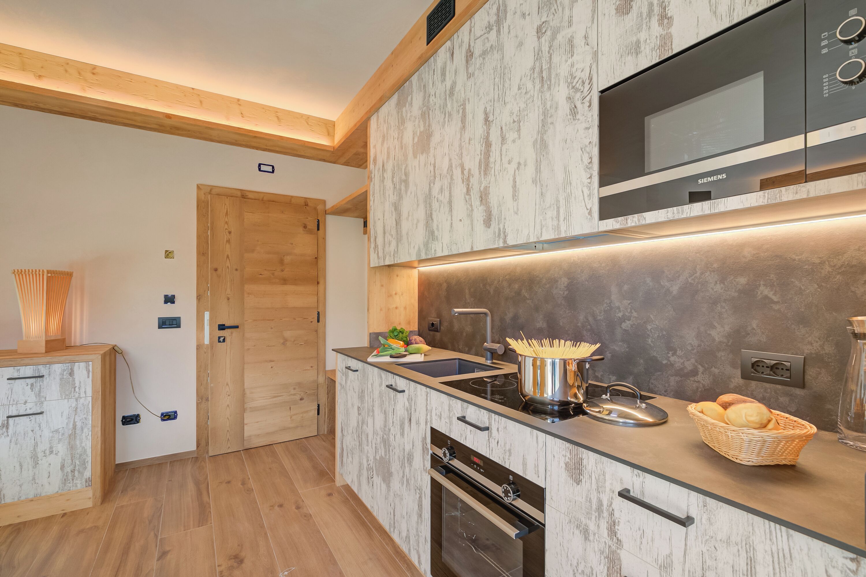 Een moderne keuken met een ingebouwde magnetron, oven en spoelbak. Er is een houten deur en een kast met lades.