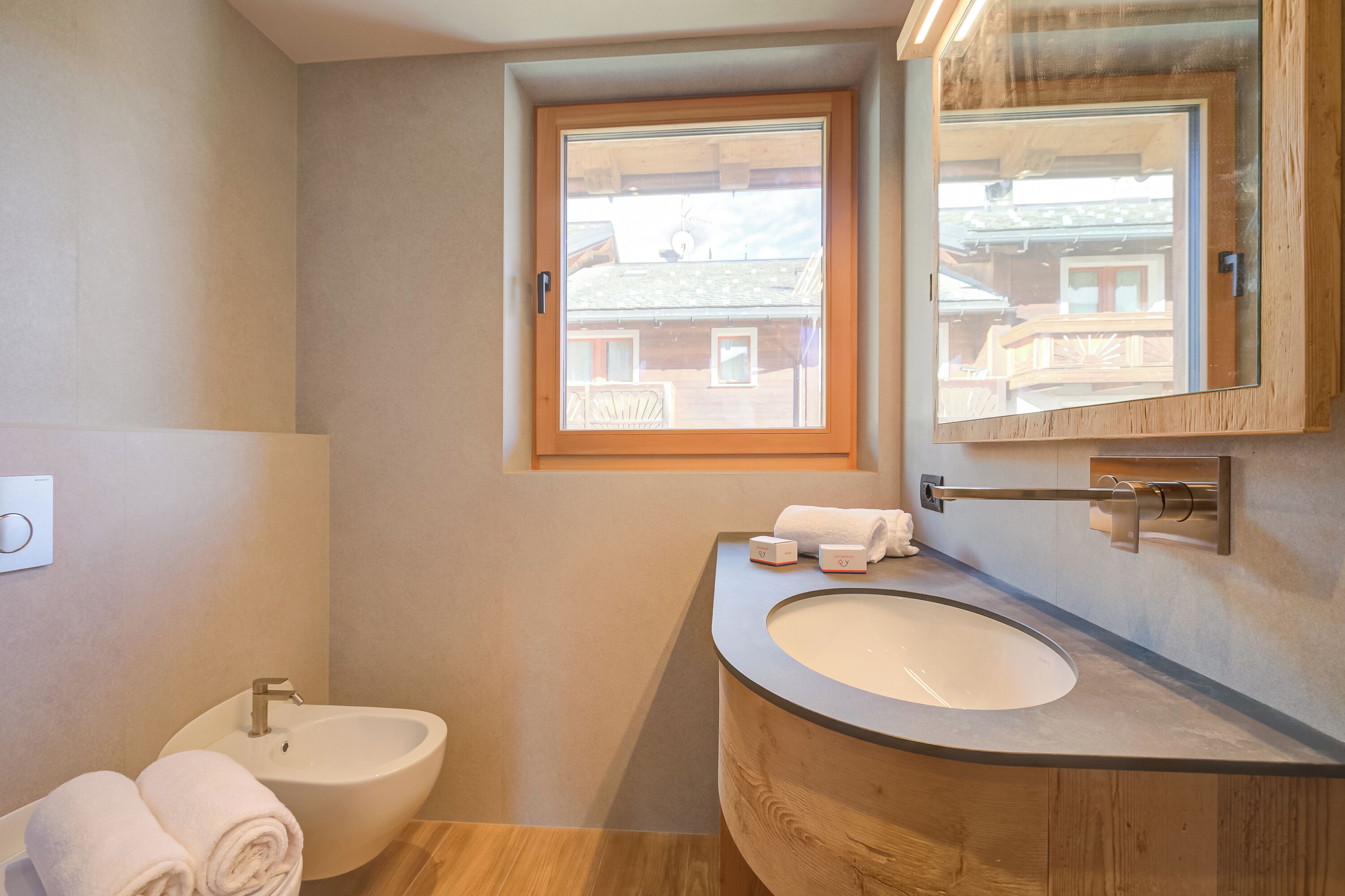 Une salle de bain avec un meuble-lavabo en bois, un lavabo, un bidet et une fenêtre donnant sur des immeubles.