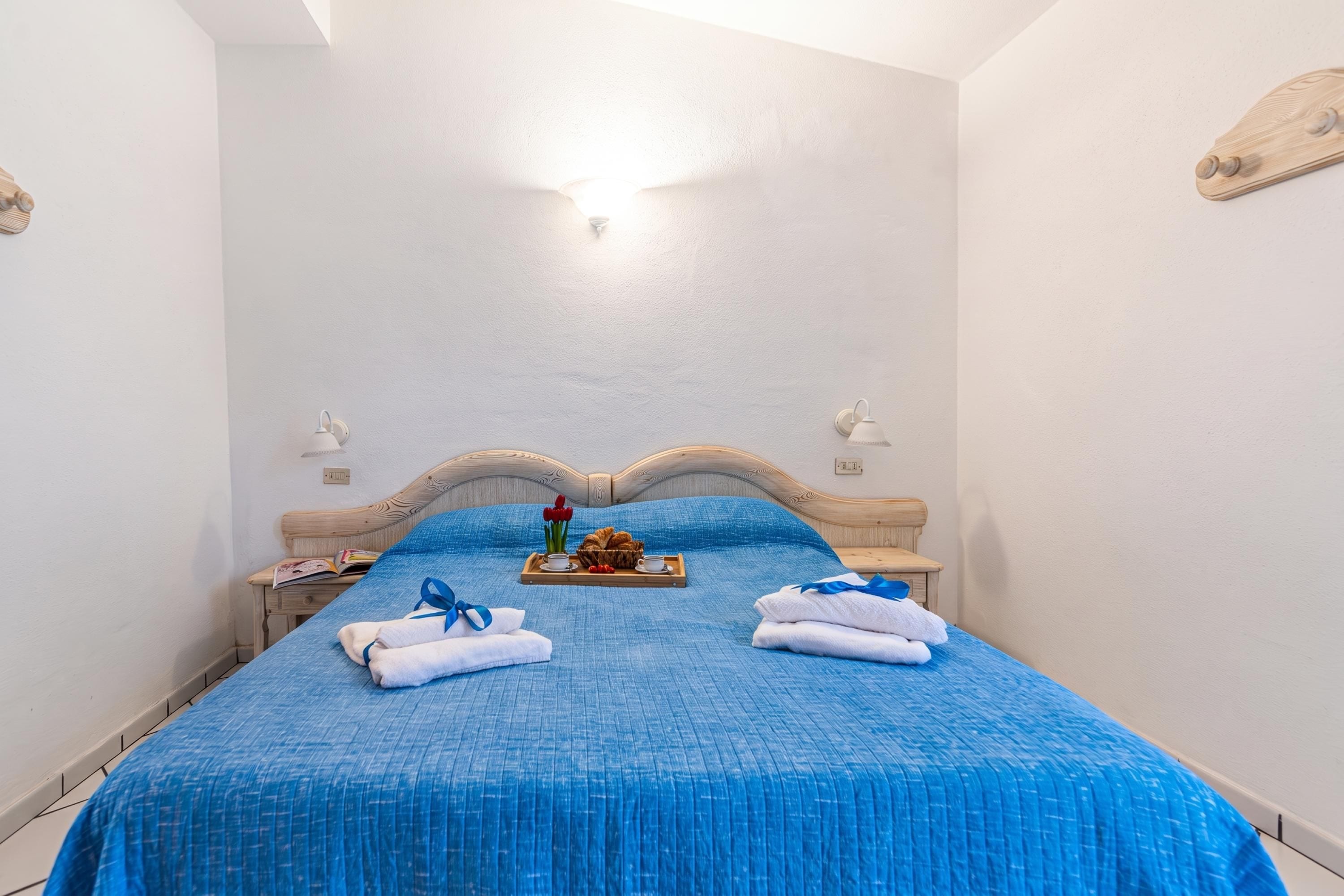 A neatly made bed with a blue bedspread, two towels, and a tray with croissants and flowers.