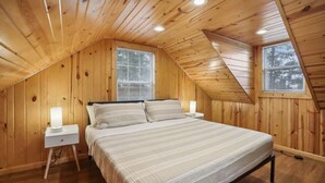A cozy attic bedroom with wooden walls, a bed with striped bedding, two bedside lamps, and two windows with blinds. - Hines Terrace Retreat - Hot Tub & Firepit (Wisconsin Dells)