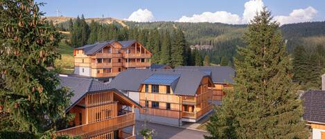 A wooden lodge with multiple balconies, surrounded by forested hills and a clear sky.