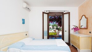 A hotel room with two beds, a dresser, a mirror, and a view of an outdoor area with tables and chairs. - Two-room Apartment in a Complex (Villasimius)