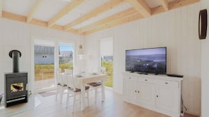 A modern kitchen with a built-in stove, a dining area with white chairs, a large TV on a white cabinet, and a view of the outdoors through a glass door.