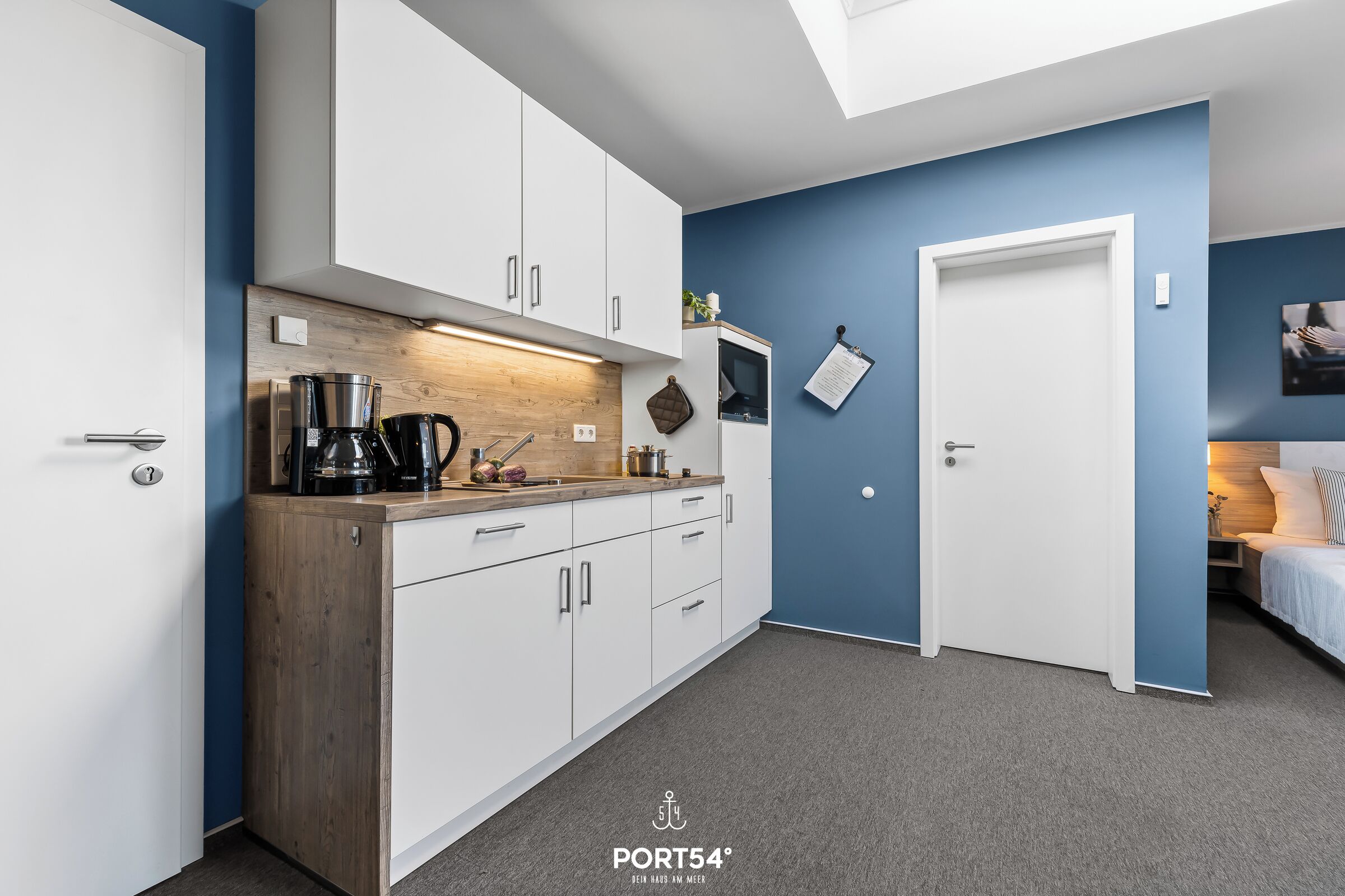 Una cocina moderna con armarios blancos, una encimera de madera y una cafetera. Se ve una puerta que da a un dormitorio con una cama y una mesita de noche.