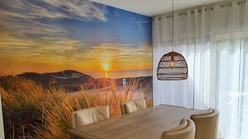 A dining area with a wooden table and chairs, a wall mural of a sunset, and a hanging light fixture.