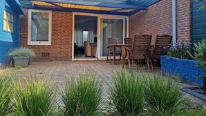 Une terrasse avec un mur en briques, une porte bleue et un espace salon abrité avec des chaises en bois.