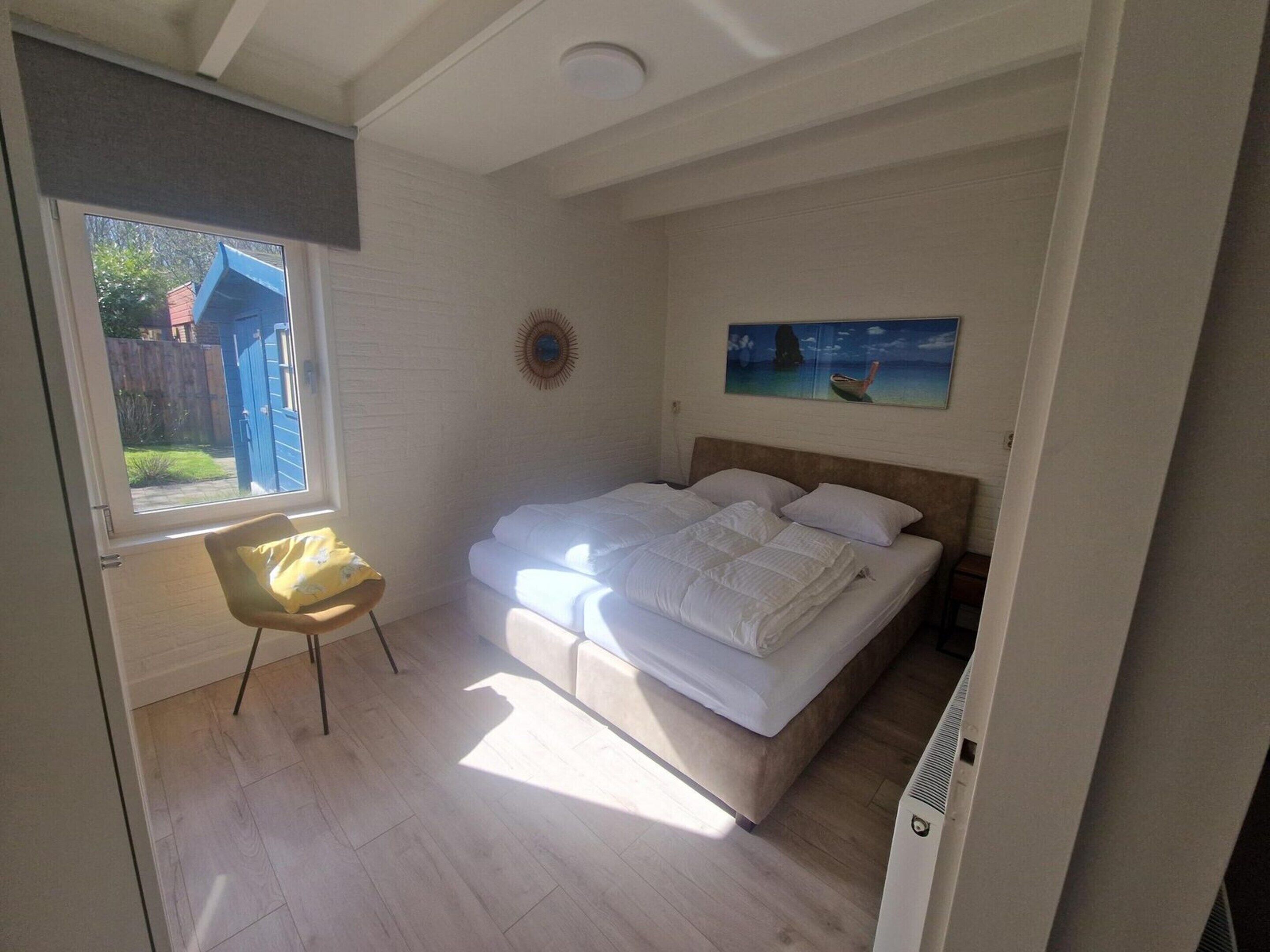 A bedroom with a bed, a chair, a painting on the wall, and a window with a view of the outside.