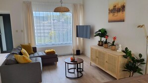 Un salon moderne avec un canapé gris, des coussins jaunes, une table basse ronde, un meuble en bois orné de plantes et une grande fenêtre avec des rideaux légers.