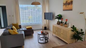 A modern living room with a grey sofa, yellow cushions, a round coffee table, a wooden cabinet with plants, and a large window with sheer curtains.
