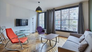 A modern living room with a sofa, a rocking chair, a table, a TV, and a large window with curtains. - Superb 3 Bedroom Flat in the Center of Paris (Paris)