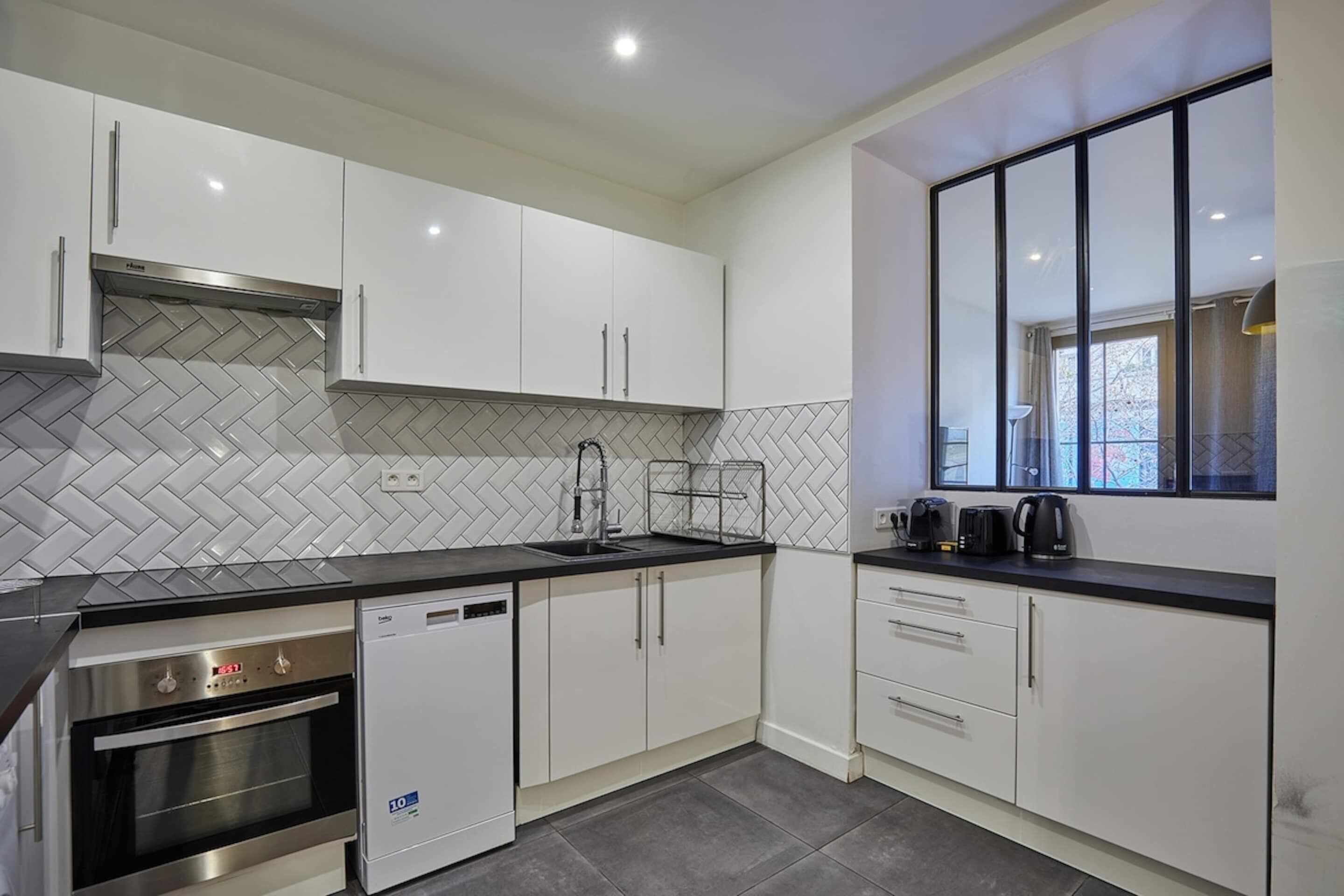 A modern kitchen with white cabinets, a herringbone tile backsplash, a dishwasher, an oven, and a window with a view.