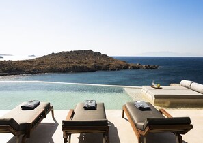 A poolside lounge area with sun loungers and a view of the sea and a rocky island. - Waterfront Villa With Private Pool (Mykonos)