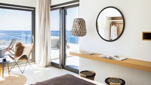 Una habitación de hotel moderna con un ventanal grande, un espejo redondo, una silla de mimbre y una estantería de madera con un libro.