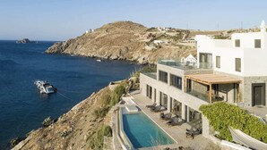 Una villa moderna con piscina, vista a una zona costera, un barco anclado cerca y un paisaje montañoso al fondo.