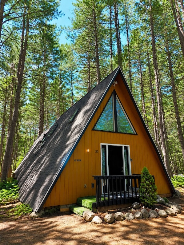 A small, triangular wooden cabin with a balcony, surrounded by a forest.