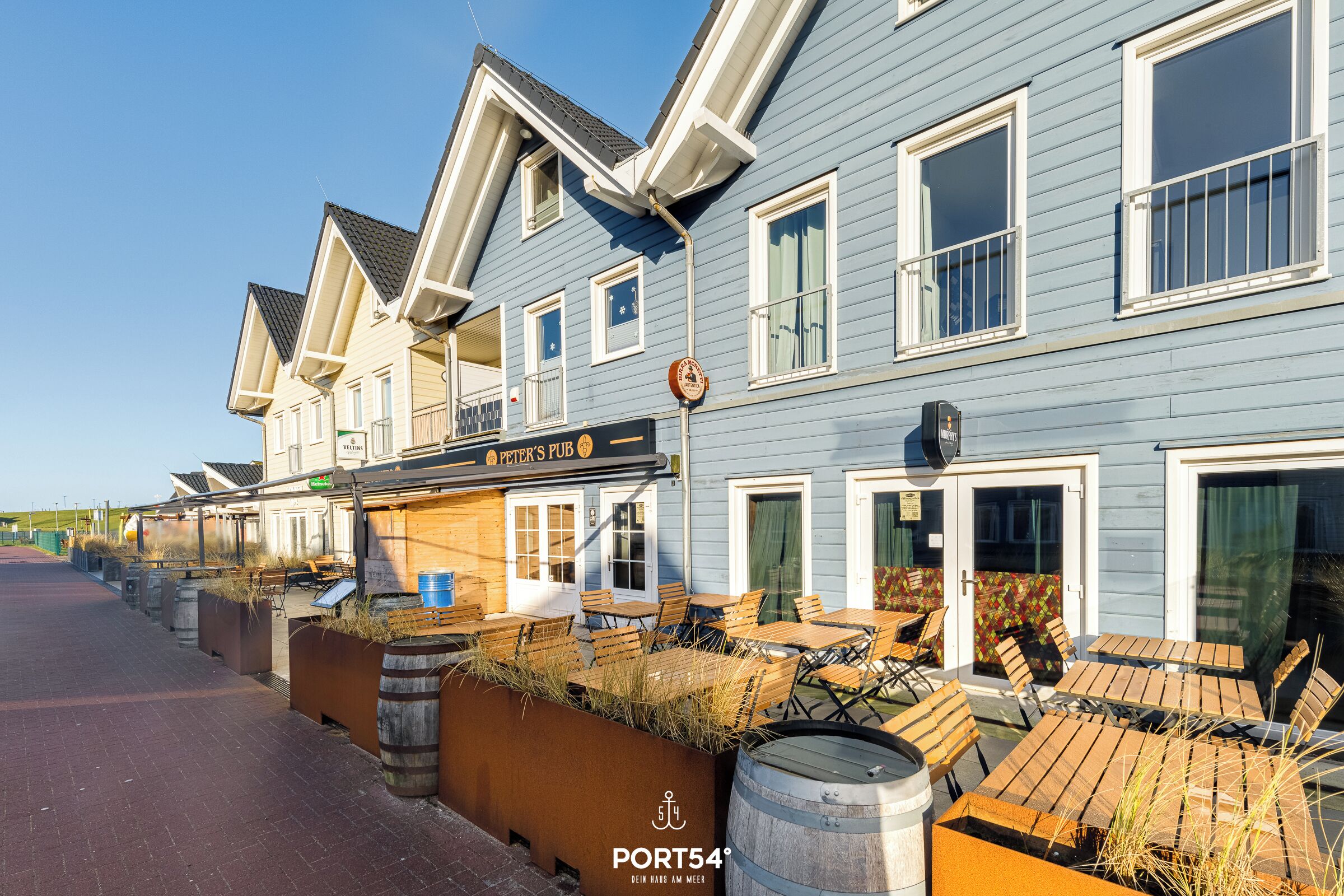 Een rij blauwe houten huizen met buiten zitplaatsen, een bord met 'Peter's Pub' erop en een vat.