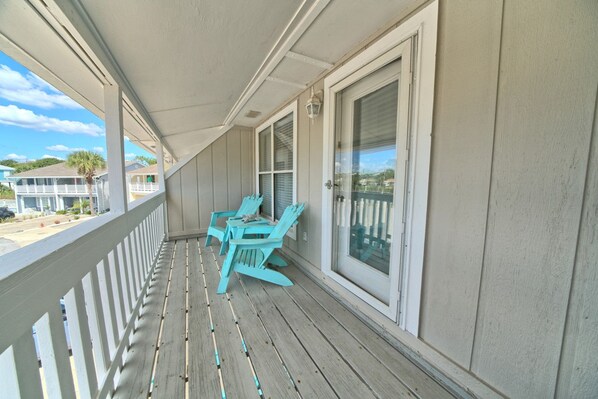 A wooden porch with a white railing, a table, and two turquoise chairs.
