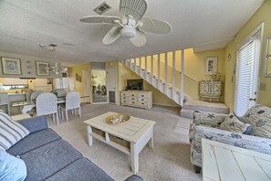 Un salon avec un canapé, une table basse, un fauteuil à motifs, un escalier et, au fond, un coin cuisine.