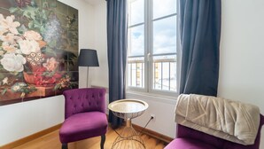 Una habitación con una pared de azulejos con motivos florales, dos sillones morados, una pequeña mesa redonda y una ventana con cortinas.