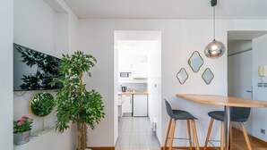 Una cocina moderna con armarios blancos, un microondas y un lavamanos. Hay una zona de comedor con una mesa de madera y dos sillas. Una gran planta en maceta está en el suelo, y un espejo cuelga de la pared.