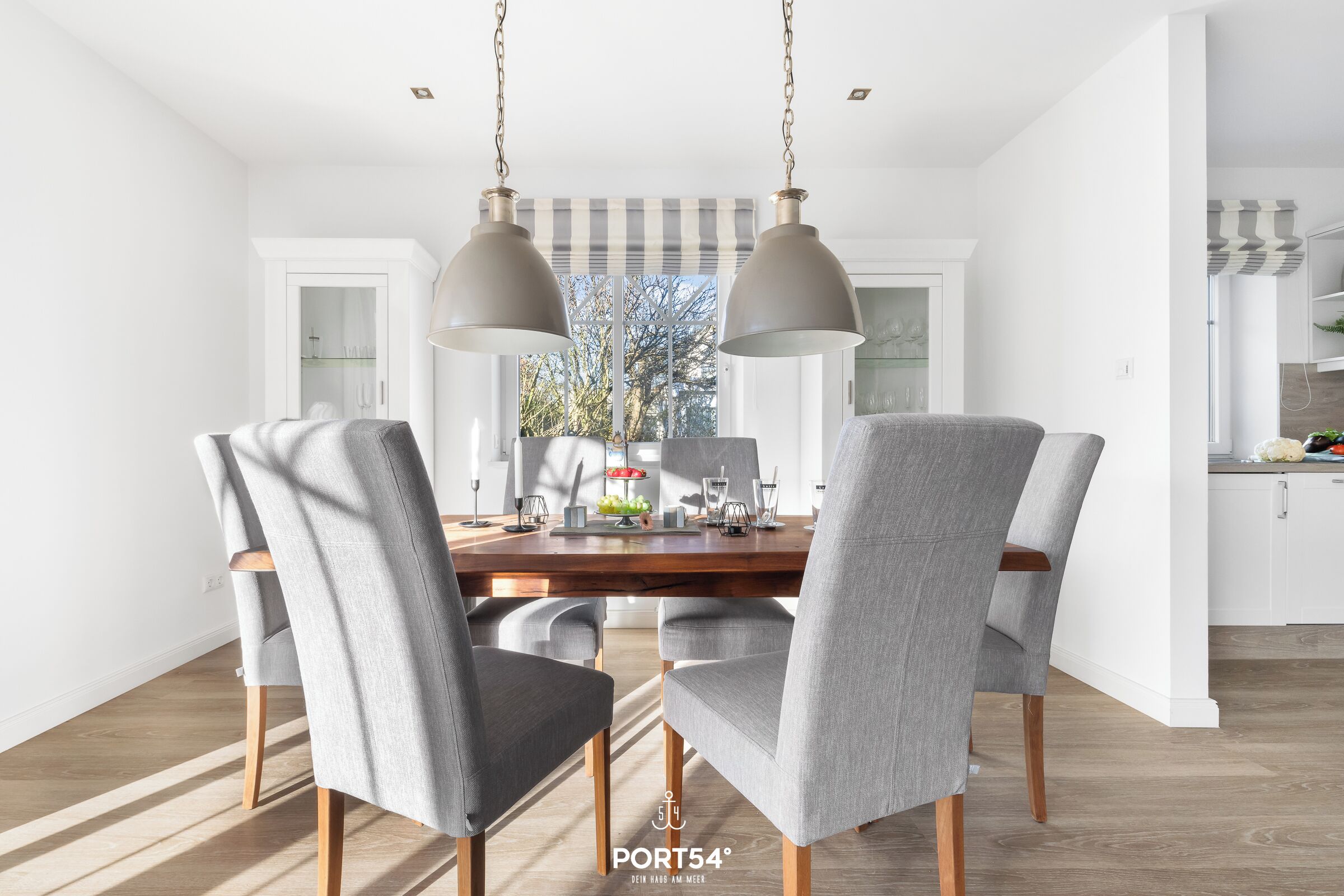 Un comedor moderno con una mesa de madera y sillas tapizadas en gris, dos lámparas colgantes y vista a la cocina al fondo.