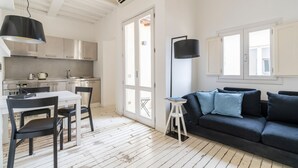 Una cocina moderna con armarios blancos y un comedor con mesa blanca y sillas negras. Hay un sofá negro con cojines azules en la sala de estar.