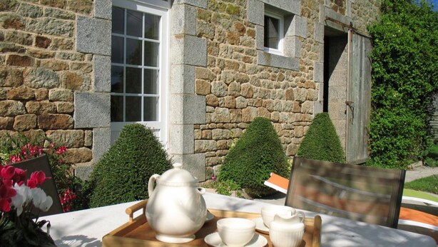 Un edificio de piedra con una ventana y una puerta, una mesa con una bandeja de tazas de té de cerámica blanca y una tetera, y arbustos bien podados.