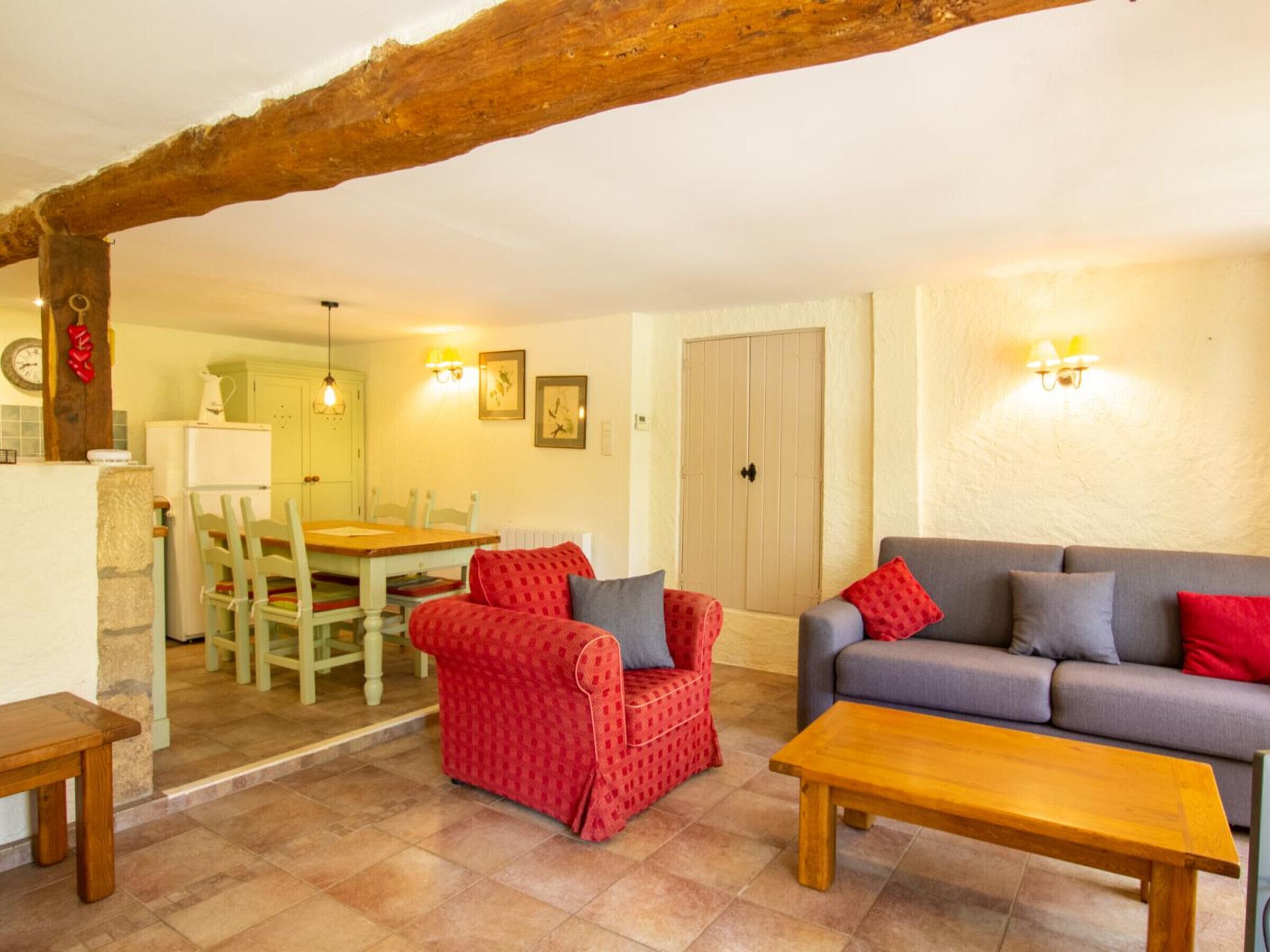 A cozy living room with a wooden table, a red patterned sofa, and a grey sofa with red cushions. There is a dining area with a wooden table and chairs, and a kitchen area with a refrigerator and a clock on the wall.